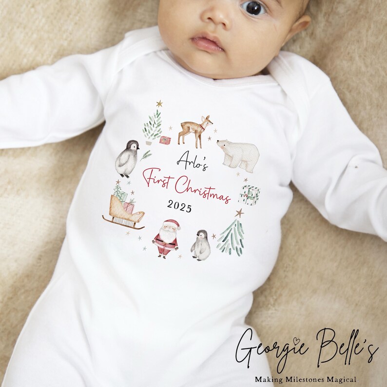 May include: A white long-sleeved baby bodysuit featuring a watercolor-style Christmas wreath graphic. The design includes a deer, polar bear, Santa Claus, penguins, trees, and a sleigh, with the text Arlo's First Christmas 2025 in the center.