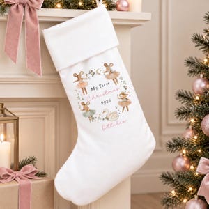 May include: A white Christmas stocking featuring a printed wreath of four mice in tutus and a swan. The center text reads My First Christmas 2026, with the name Ottilie printed in pink below. The stocking has a wide white cuff at the top.