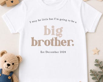 May include: A white short-sleeved t-shirt featuring the text 'I may be little but I'm going to be a big brother.' in black and beige lettering, with 'Est December 2024' printed below. The shirt is surrounded by a teddy bear and gift tags.