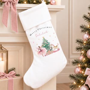 May include: A white Christmas stocking featuring a folded cuff and a printed graphic of a ballerina in a pink tutu, a decorated Christmas tree, and a rocking horse. The text Merry Christmas and the name Delilah are printed in black and pink.