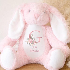 Puede incluir: Un peluche rosa con forma de conejo, con orejas largas y el interior blanco. El conejo tiene un &oacute;valo blanco en el pecho con la letra "G" y el nombre "Gracie", rodeado de detalles florales.