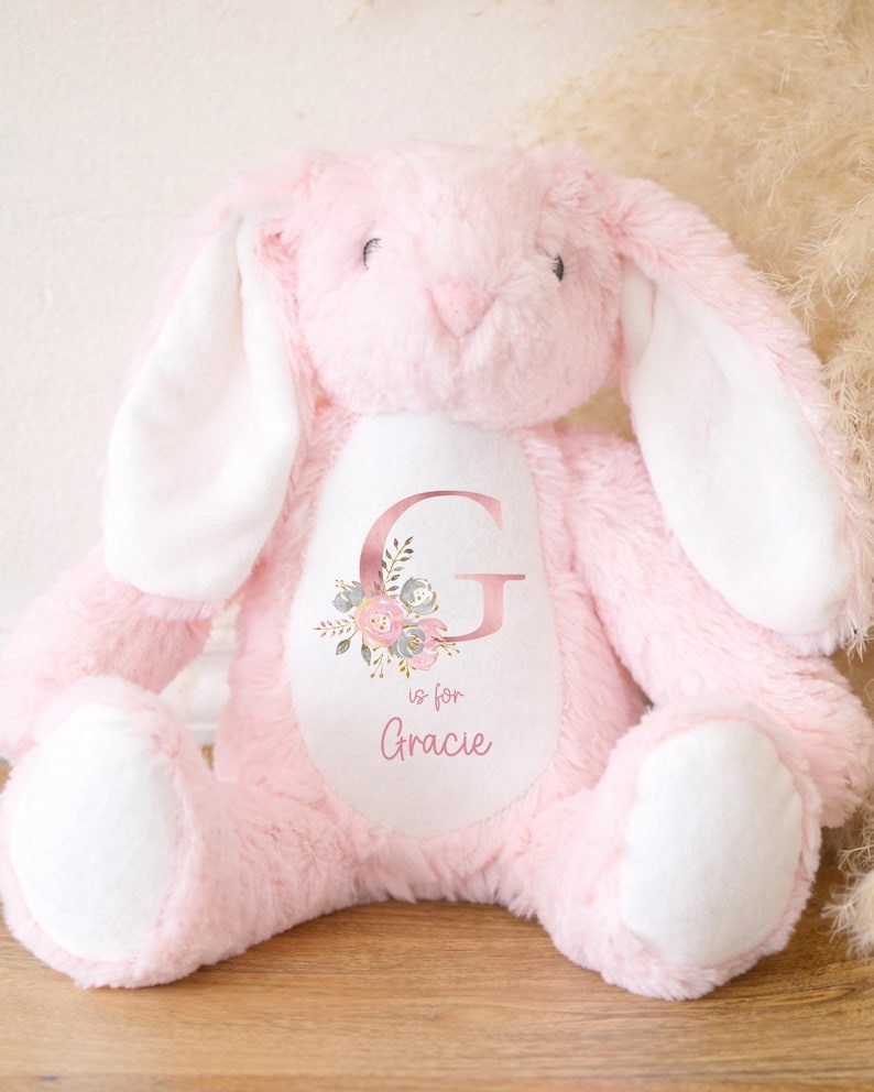 Puede incluir: Un peluche rosa con forma de conejo, con orejas largas y el interior blanco. El conejo tiene un &oacute;valo blanco en el pecho con la letra "G" y el nombre "Gracie", rodeado de detalles florales.
