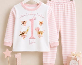 May include: A white long-sleeve shirt with pink striped sleeves and matching pink striped pants. The shirt features the text "When I wake up I will be" above a large pink number "1" and the name "Lyla," surrounded by illustrations of four fairies.