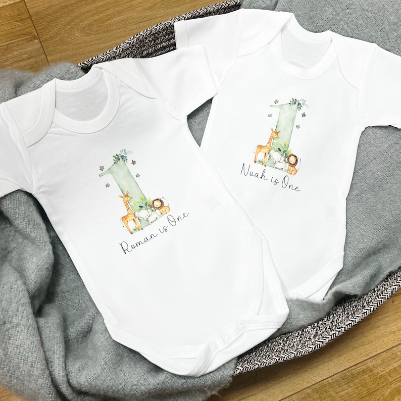 May include: Two white baby onesies with a green number one, a giraffe, a lion, and a white animal. The onesies say "Roman is One" and "Noah is One".