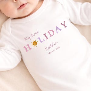 May include: White baby onesie with long sleeves. The onesie features the text "My First HOLIDAY" in purple and pink, a smiling sun graphic, the name "Hallie", and "Majorca 2026".