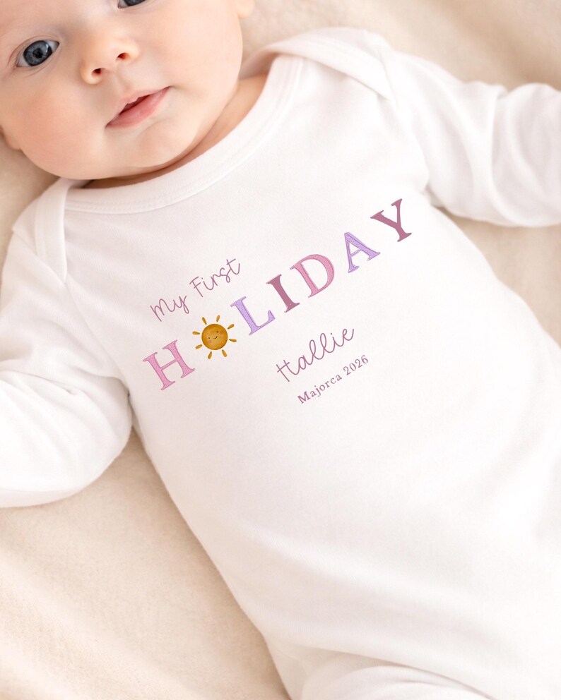 May include: White baby onesie with long sleeves. The onesie features the text "My First HOLIDAY" in purple and pink, a smiling sun graphic, the name "Hallie", and "Majorca 2026".