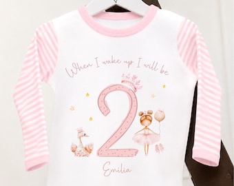 May include: White long-sleeved shirt with pink striped sleeves and a pink neckline. The shirt features a large pink number 2 with a ballerina, swan, and the name "Emilia". Text reads "When I wake up I will be".