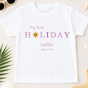 May include: White t-shirt with the text "My First HOLIDAY" in pink and purple, featuring a sun graphic. The name "Hallie" and "Majorca 2026" are printed below. The shirt is surrounded by beach-themed accessories.