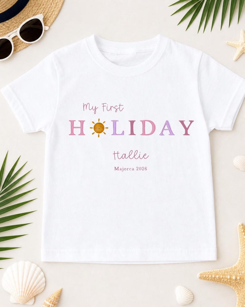 May include: White t-shirt with the text "My First HOLIDAY" in pink and purple, featuring a sun graphic. The name "Hallie" and "Majorca 2026" are printed below. The shirt is surrounded by beach-themed accessories.
