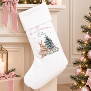 May include: A white Christmas stocking featuring a printed illustration of a bunny wearing a tutu next to a decorated Christmas tree and wrapped gifts. The text Merry Christmas and the name Callie are printed in pink and black script above the bunny.