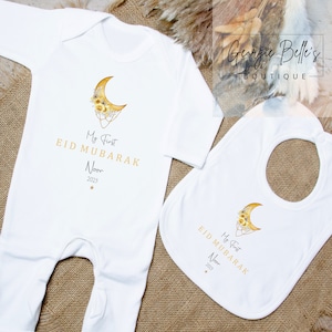 Puede incluir: Mono blanco para beb&eacute; y babero con una luna creciente dorada y un dise&ntilde;o floral. El texto "My First Eid Mubarak Noor 2023" est&aacute; impreso en ambos art&iacute;culos.