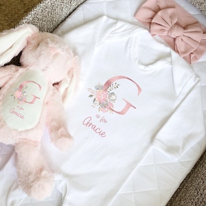 Puede incluir: Body de beb&eacute; blanco con una gran letra "G" en oro rosa y dise&ntilde;o floral. El texto "is for Gracie" est&aacute; debajo. Un conejito de peluche rosa y una diadema rosa con lazo tambi&eacute;n est&aacute;n en la imagen.
