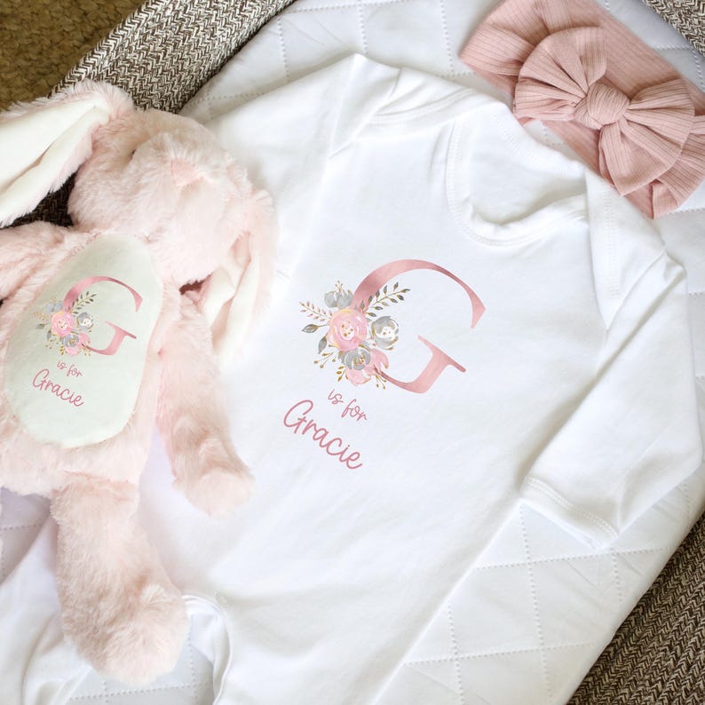 Puede incluir: Body de beb&eacute; blanco con una gran letra "G" en oro rosa y dise&ntilde;o floral. El texto "is for Gracie" est&aacute; debajo. Un conejito de peluche rosa y una diadema rosa con lazo tambi&eacute;n est&aacute;n en la imagen.