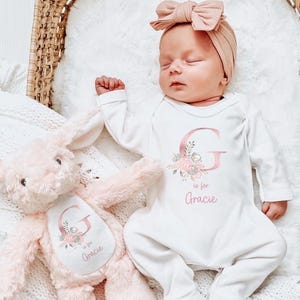 Puede incluir: Un beb&eacute; dormido con un body blanco con una gran letra rosa "G" y el nombre "Gracie". Una diadema rosa a juego y un conejito de peluche rosa tambi&eacute;n est&aacute;n en la imagen. El body y el conejito tienen detalles florales.