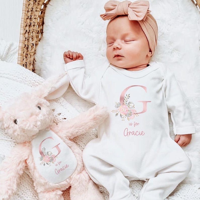 Puede incluir: Un beb&eacute; dormido con un body blanco con una gran letra rosa "G" y el nombre "Gracie". Una diadema rosa a juego y un conejito de peluche rosa tambi&eacute;n est&aacute;n en la imagen. El body y el conejito tienen detalles florales.