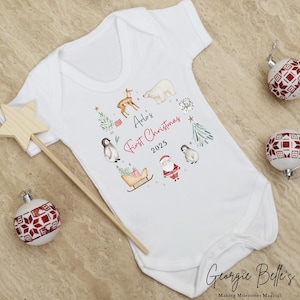 May include: A white short-sleeved baby bodysuit featuring a festive watercolor print of a polar bear, reindeer, penguins, Santa Claus, and a sleigh. The text reads Arlo's First Christmas 2025 in black and red script and block lettering.