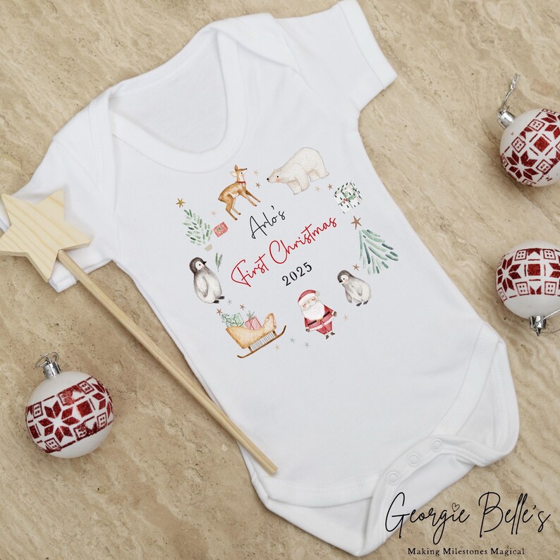 May include: A white short-sleeved baby bodysuit featuring a festive watercolor print of a polar bear, reindeer, penguins, Santa Claus, and a sleigh. The text reads Arlo's First Christmas 2025 in black and red script and block lettering.