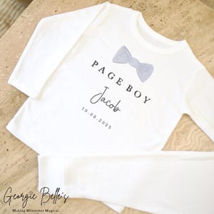 May include: White long-sleeved pajama set featuring the text "PAGE BOY" above a light blue bow tie graphic. The name "Jacob" and the date "10.08.2025" are printed below. The brand name "Georgie Belle's" is at the bottom.
