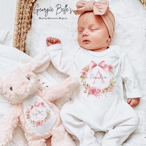 May include: A baby in a white onesie featuring a floral wreath design and the name "Amelia". The image also includes a pink bow headband and a pink plush bunny. The text "Georgie Belle's" is visible at the top of the image.