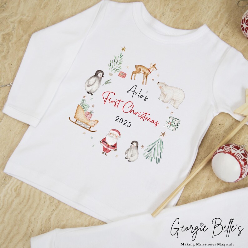 May include: A white long-sleeve shirt featuring a watercolor-style Christmas graphic with a deer, polar bear, penguins, Santa Claus, a sleigh, and gifts. The text reads Arlo's First Christmas 2025 in black and red script and print lettering.
