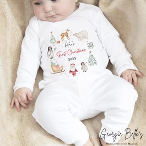 May include: A white long-sleeved baby outfit featuring a festive graphic with a deer, polar bear, penguins, Santa Claus, a sleigh, and Christmas trees. The text reads Arlo's First Christmas 2025 in black and red script and block lettering.