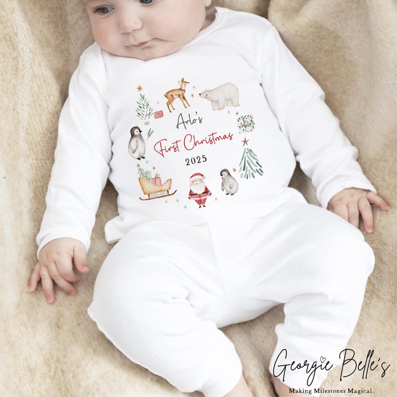 May include: A white long-sleeved baby outfit featuring a festive graphic with a deer, polar bear, penguins, Santa Claus, a sleigh, and Christmas trees. The text reads Arlo's First Christmas 2025 in black and red script and block lettering.