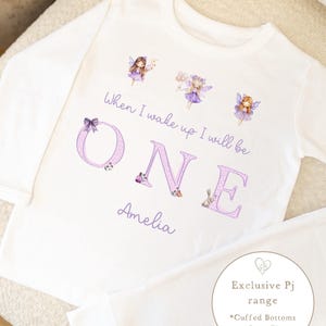 May include: White pyjama set with long sleeves and matching trousers. The top displays the text "When I wake up I will be ONE Amelia" in purple, with fairy illustrations. A round graphic with text is also included.