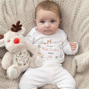 May include: A white long-sleeve shirt and pants set featuring a watercolor Christmas print with the text Grayson's First Christmas 2025, paired with a plush tan reindeer toy that displays the same matching holiday graphic and text.