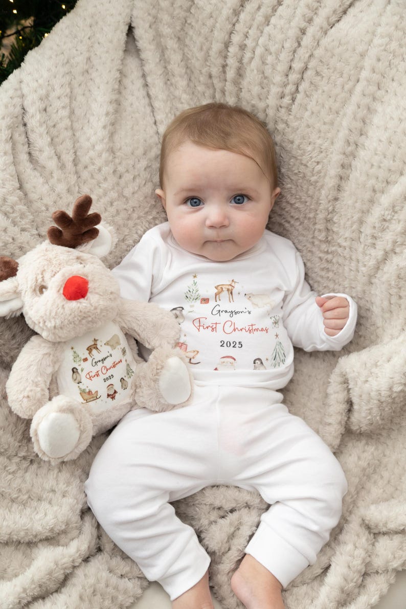May include: A white long-sleeve shirt and pants set featuring a watercolor Christmas print with the text Grayson's First Christmas 2025, paired with a plush tan reindeer toy that displays the same matching holiday graphic and text.