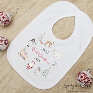 May include: A white baby bib featuring a watercolor illustration of a penguin, a deer, a polar bear, a sleigh, and Santa Claus. The text reads Arlo's First Christmas 2025 in red and black script and print lettering, surrounded by small green trees.