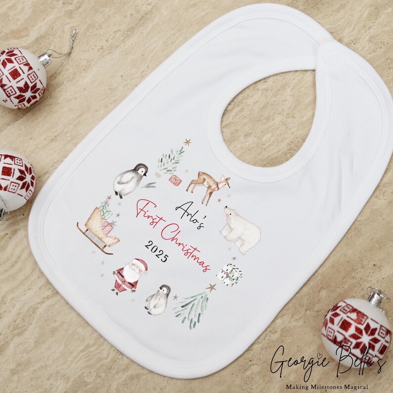 May include: A white baby bib featuring a watercolor illustration of a penguin, a deer, a polar bear, a sleigh, and Santa Claus. The text reads Arlo's First Christmas 2025 in red and black script and print lettering, surrounded by small green trees.
