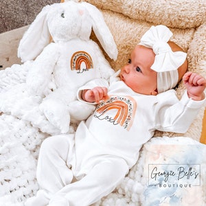 Puede incluir: Un body blanco para bebé con un diseño de arcoíris y flores y el nombre "Lola" impreso en él. El body lo lleva un bebé que lleva una diadema blanca. Un conejito de peluche blanco con un diseño de arcoíris y flores y el nombre "Lola" impreso en él está en el fondo.
