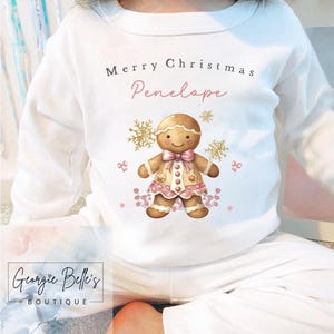 May include: White long-sleeved pajama top with a gingerbread person graphic. The graphic includes the text "Merry Christmas Penelope" in black and pink script. The gingerbread person wears a pink dress and bow.