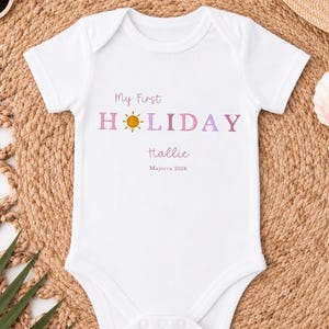 May include: White baby bodysuit with the text "My First Holiday Hallie Mallorca 2026" in pink and purple. The word "Holiday" has a sun graphic. The bodysuit is short-sleeved and has a round neckline.