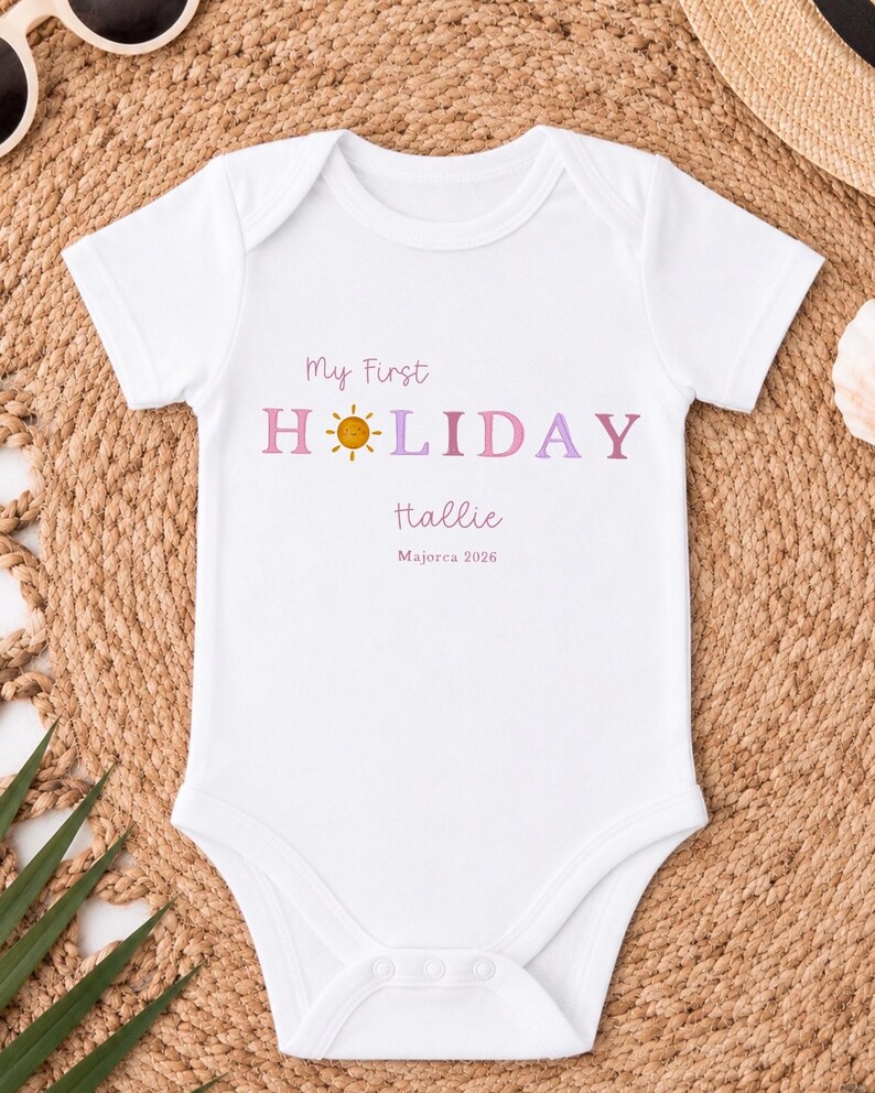 May include: White baby bodysuit with the text "My First Holiday Hallie Mallorca 2026" in pink and purple. The word "Holiday" has a sun graphic. The bodysuit is short-sleeved and has a round neckline.