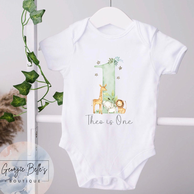 May include: White baby onesie with a green number one, a giraffe, an elephant, and a lion. The text "Theo is One" is printed below the animals.