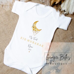 Puede incluir: Un body blanco para beb&eacute; con una luna creciente dorada y un dise&ntilde;o floral. El texto "My First Eid Mubarak Noor 2023" est&aacute; impreso en el body.