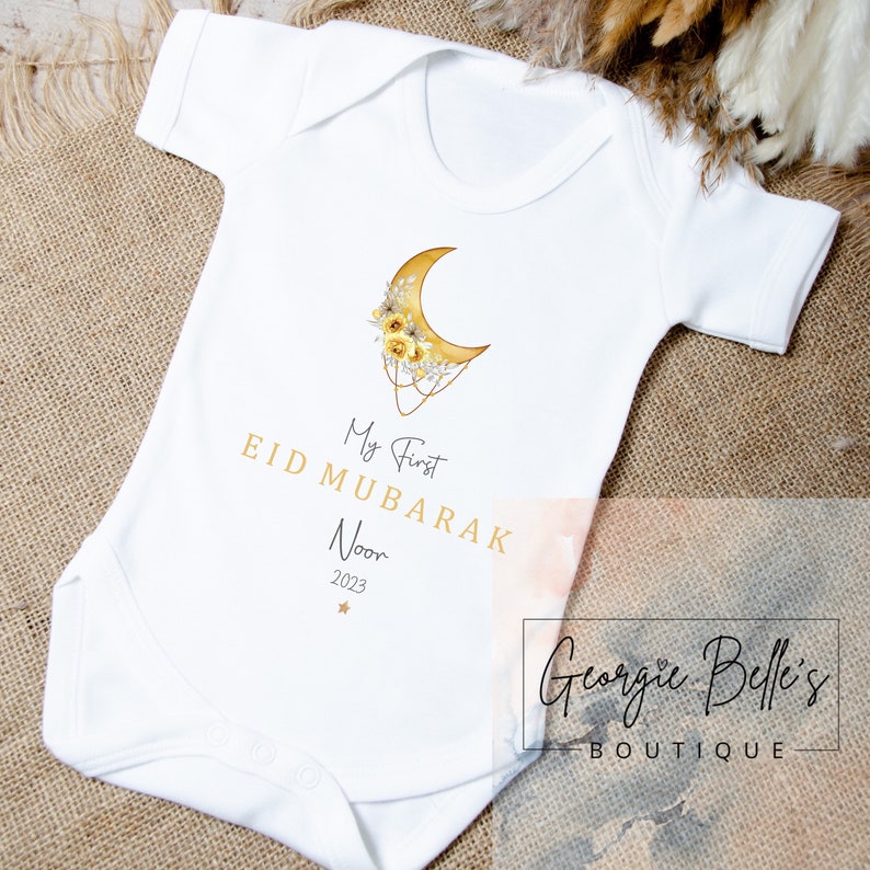 Puede incluir: Un body blanco para beb&eacute; con una luna creciente dorada y un dise&ntilde;o floral. El texto "My First Eid Mubarak Noor 2023" est&aacute; impreso en el body.