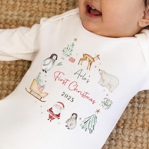 May include: A white long-sleeve baby bodysuit featuring a watercolor-style circular print of a deer, polar bear, penguins, Santa Claus, a sleigh, and gifts. The text reads Arlo's First Christmas 2025 in black and red script and print.