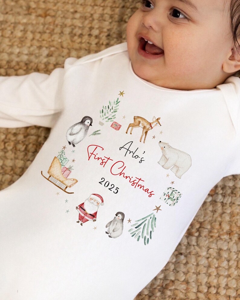 May include: A white long-sleeve baby bodysuit featuring a watercolor-style circular print of a deer, polar bear, penguins, Santa Claus, a sleigh, and gifts. The text reads Arlo's First Christmas 2025 in black and red script and print.