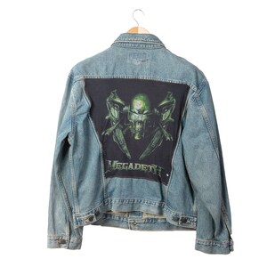 May include: A light blue denim jacket with a large black patch on the back. The patch features a green skull with crossed swords and the band name "MEGADETH" in white lettering. The jacket is hanging on a wooden hanger.