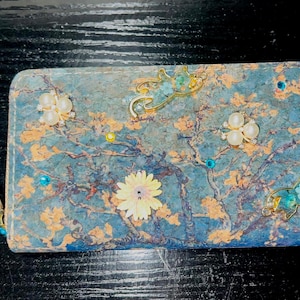 May include: A blue wallet with a floral design, adorned with pearl and jewel accents. The wallet features a tree branch pattern with yellow and gold accents, and decorative gold and turquoise elephant charms.
