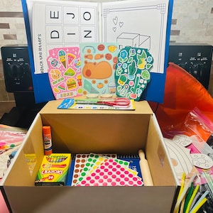 May include: A craft box filled with art supplies, including crayons, stickers, glue, and a small rolling pin. Colorful sticker sheets and cut-out shapes are visible. A blue folder with educational materials is in the background.