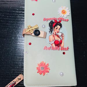 May include: A light green wallet with a decorative design. The wallet features a pin-up girl illustration with the text "Expensive difficult And talks back". Embellishments include faux pearls, red gems, and floral accents. A gold bow clasp secures the wallet.