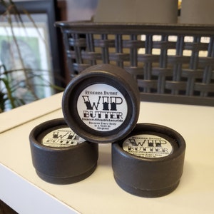 May include: Three small black containers with white labels that read "Process Butter WHIP BUTTER" and "Because Every Body is a Work in Progress".