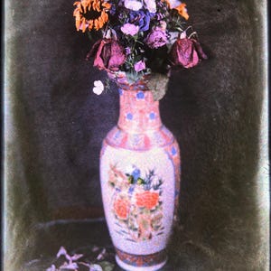 May include: A tall, slender ceramic vase with a pale pink base, decorated with blue geometric patterns and orange floral motifs, containing a bouquet of dried flowers in shades of deep purple, orange, and pink, with fallen petals scattered below.