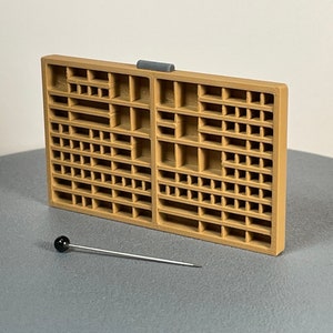 May include: A tan-colored plastic storage box with a grid of compartments. The box has a small gray latch on the top. A black pin with a black ball head is lying on a gray surface.