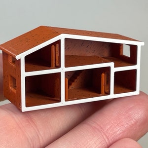 Puede incluir: Un pequeño modelo de casa impreso en 3D, de color marrón con molduras blancas. La casa está cortada para mostrar el interior, que incluye una escalera y varias habitaciones.