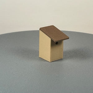 Puede incluir: Una pequeña casita de pájaros beige con un techo marrón. La casita de pájaros tiene una pequeña abertura redonda para que los pájaros puedan entrar.