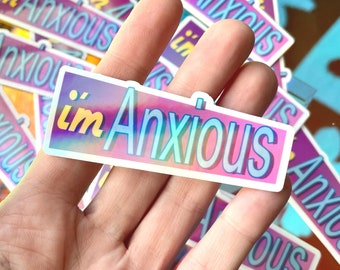 Peut inclure: Un autocollant holographique avec le texte "i'm Anxious" dans un schéma de couleurs rose, bleu et jaune. L'autocollant est tenu par une main.